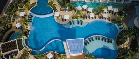 Vista aérea de un complejo turístico con una amplia piscina central, numerosas tumbonas y un edificio con balcones.