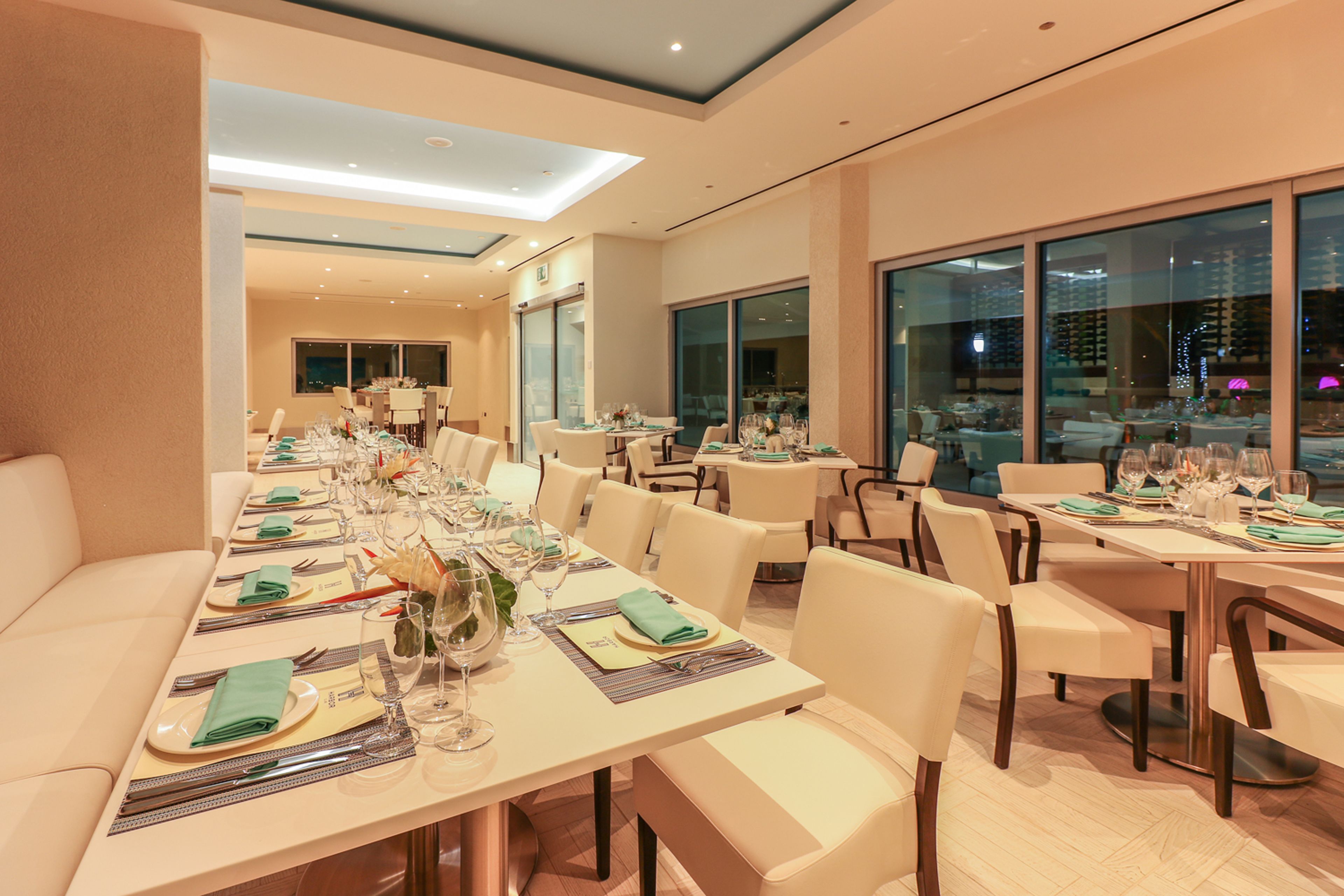 A modern dining area with long tables set for multiple guests, featuring a neutral color palette and contemporary furnishings.