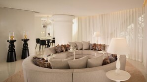 A modern living room with a curved sofa, a round coffee table, and a dining area in the background. - Deluxe Apartments (Drama)