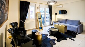 A modern living room with a grey sofa, a black coffee table, a round black ottoman, a black leather rug, and a black chair. There is a large mirror on the wall, a black floor lamp, and a flat-screen TV mounted on the wall. - Deluxe Apartments (Drama)