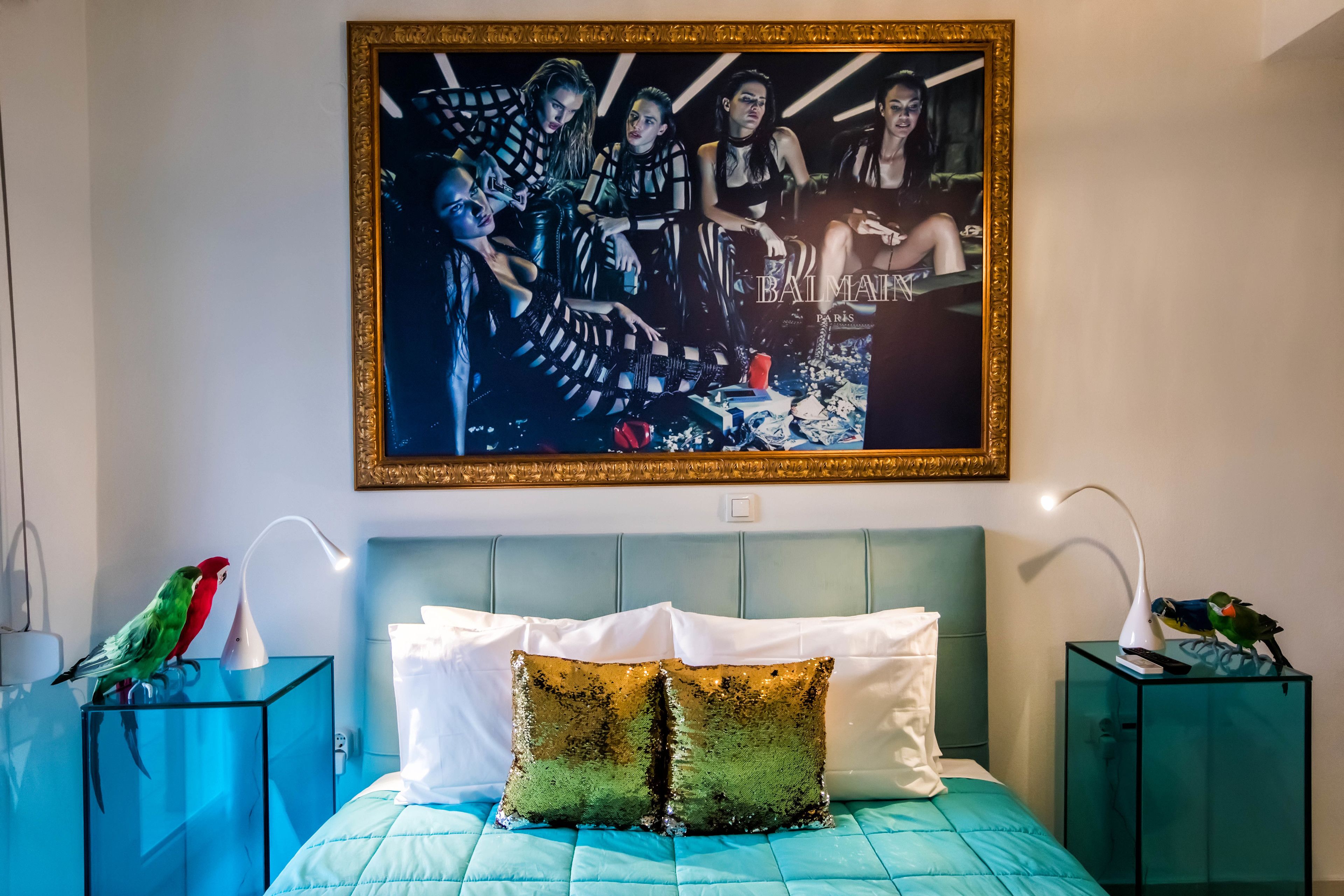 A bedroom with a teal bedspread, white pillows, a framed artwork above the bed, and two bedside tables with lamps and a parrot on each.