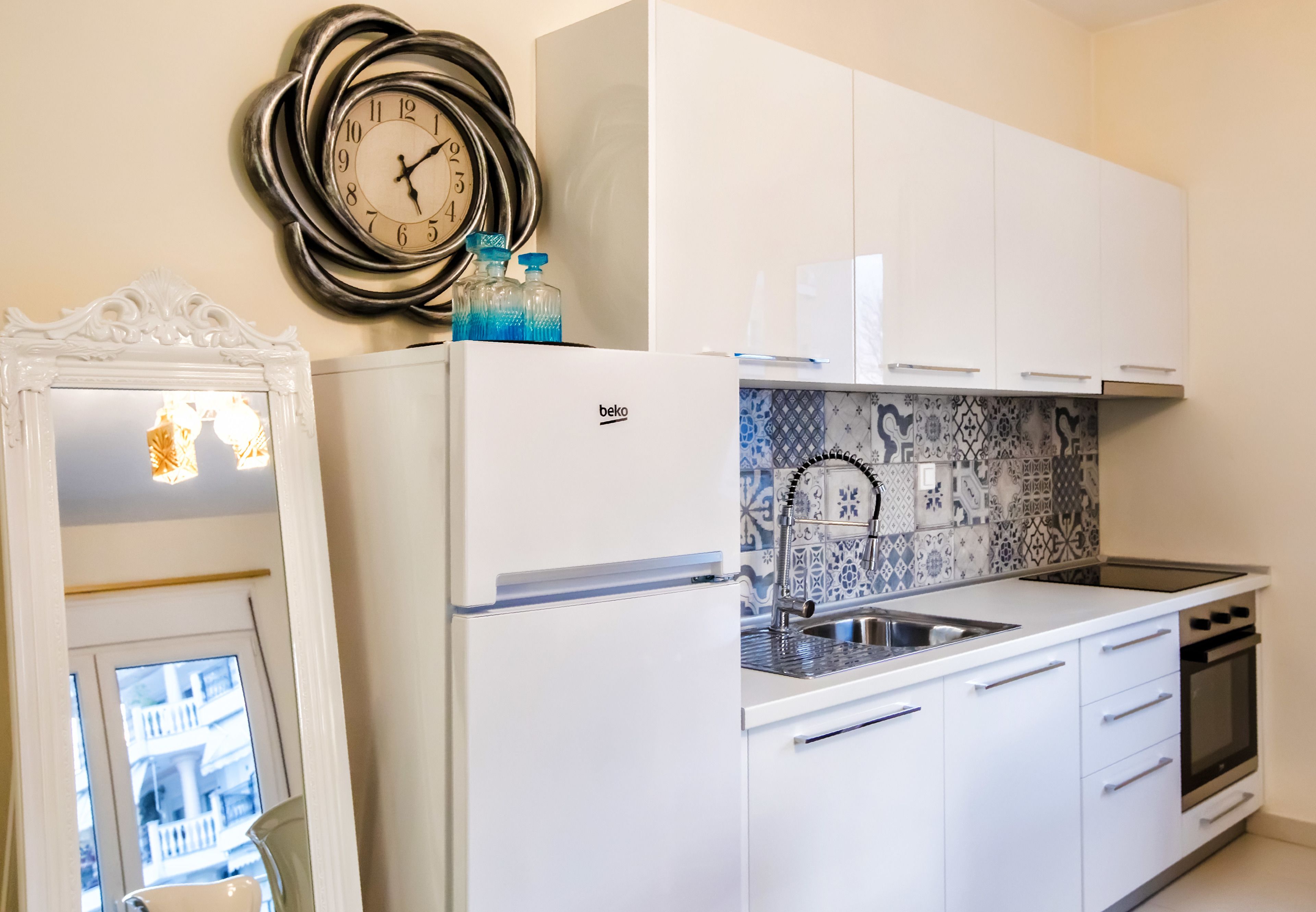 A compact kitchen with white cabinetry, a built-in sink, and a stove. A decorative clock and a mirror are mounted on the wall.