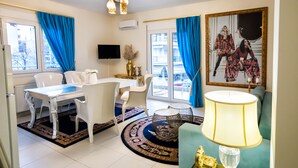A modern living room with a dining area, featuring a TV, a sofa, a table, and chairs. - Deluxe Apartments (Drama)