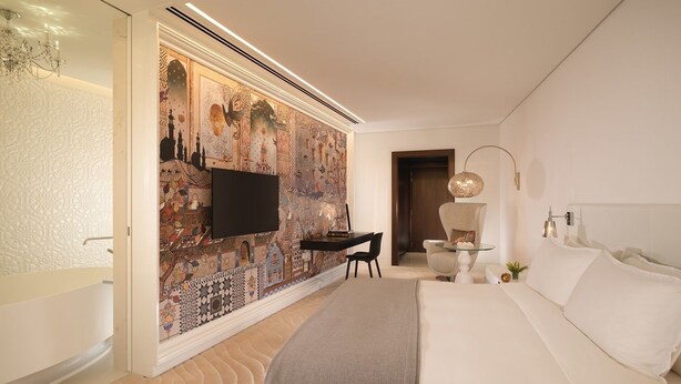 A modern hotel room with a large bed, a desk, a chair, and a television mounted on a patterned wall. - Deluxe Apartments (Drama)