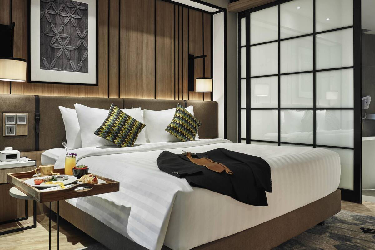 Una habitación de hotel moderna con una cama grande, una mesita de noche con una bandeja de desayuno, un ventanal amplio y una obra de arte colgada en la pared.