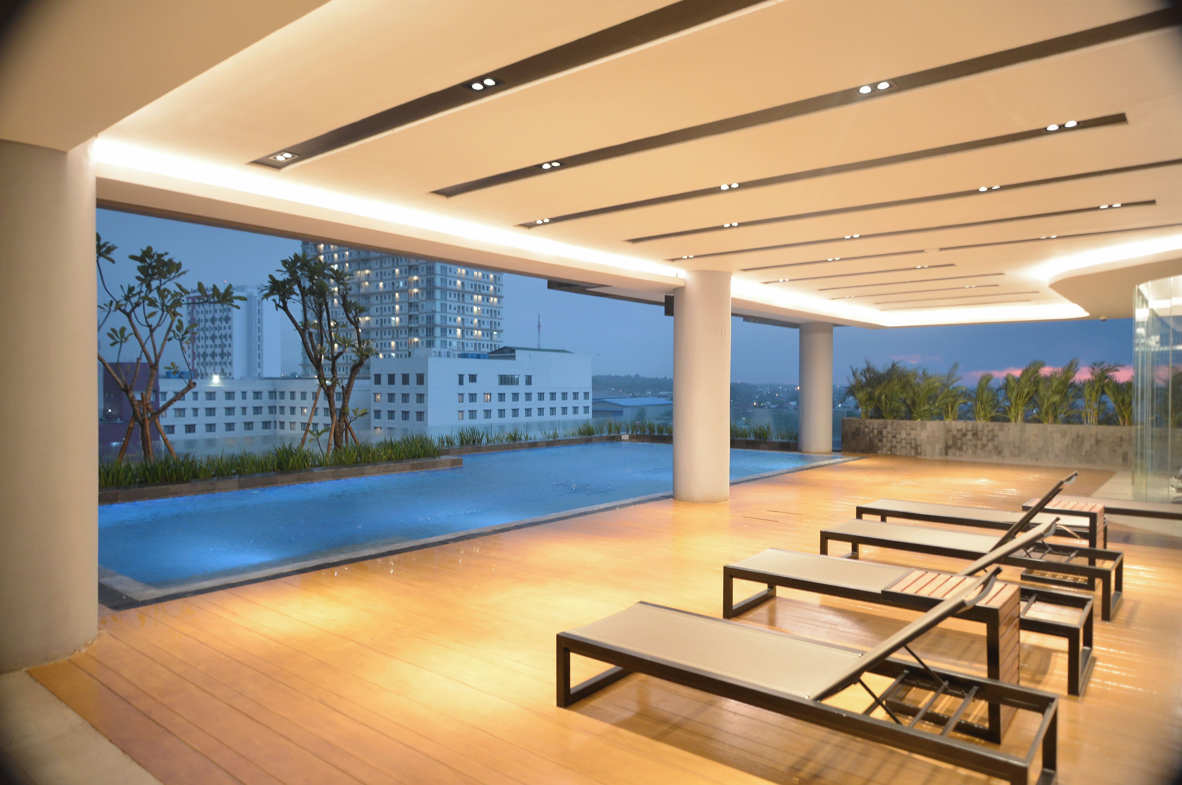 Un espace de détente extérieur moderne, doté d’une piscine, de chaises longues et offrant une vue sur le paysage urbain au crépuscule.