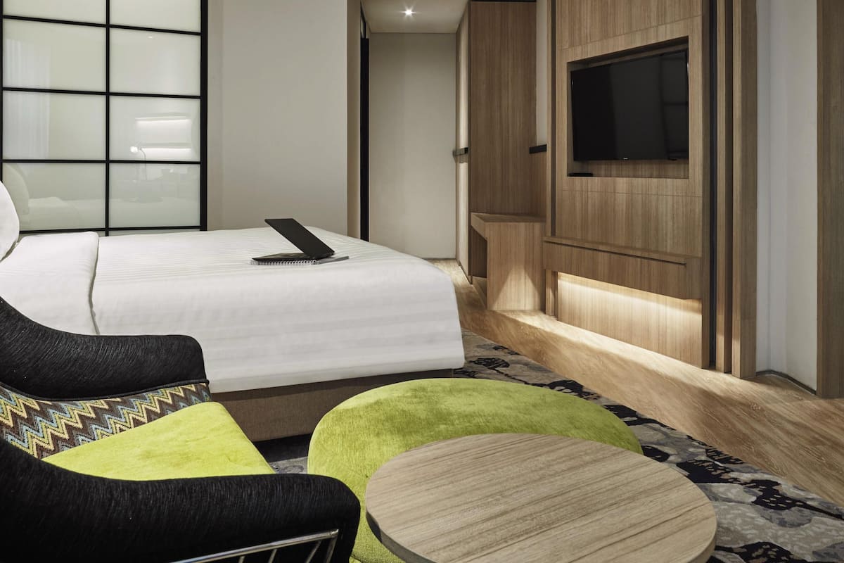 Una habitación de hotel moderna con una cama bien hecha, un televisor de pantalla plana montado en un panel de madera, una mesita redonda y una zona de estar cómoda con un tapete estampado.