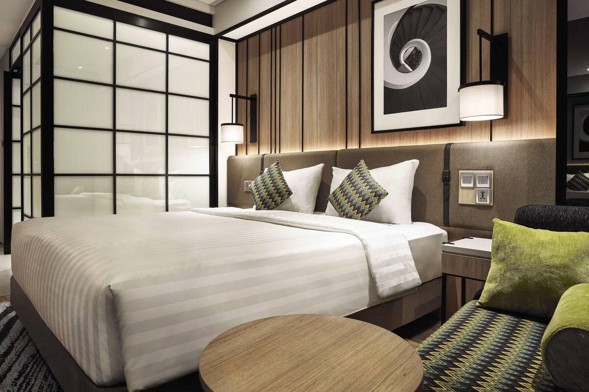 Una habitación de hotel moderna con una cama grande, una mesa redonda de madera y vistas de la ciudad a través de ventanales de gran altura.