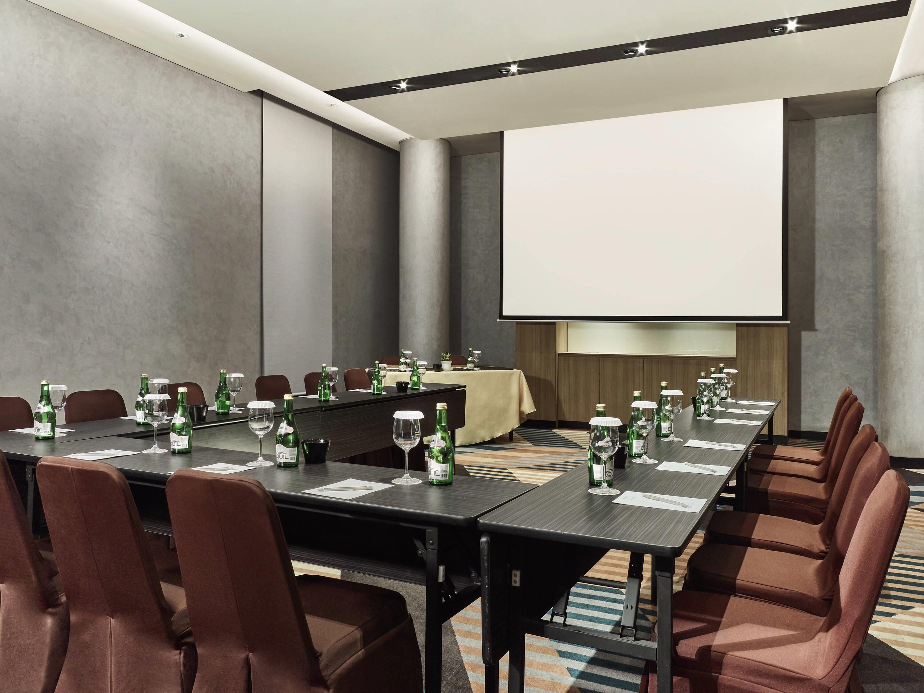 Ein Konferenzraum mit einem langen Tisch, braunen Stühlen und einer Projektorleinwand.