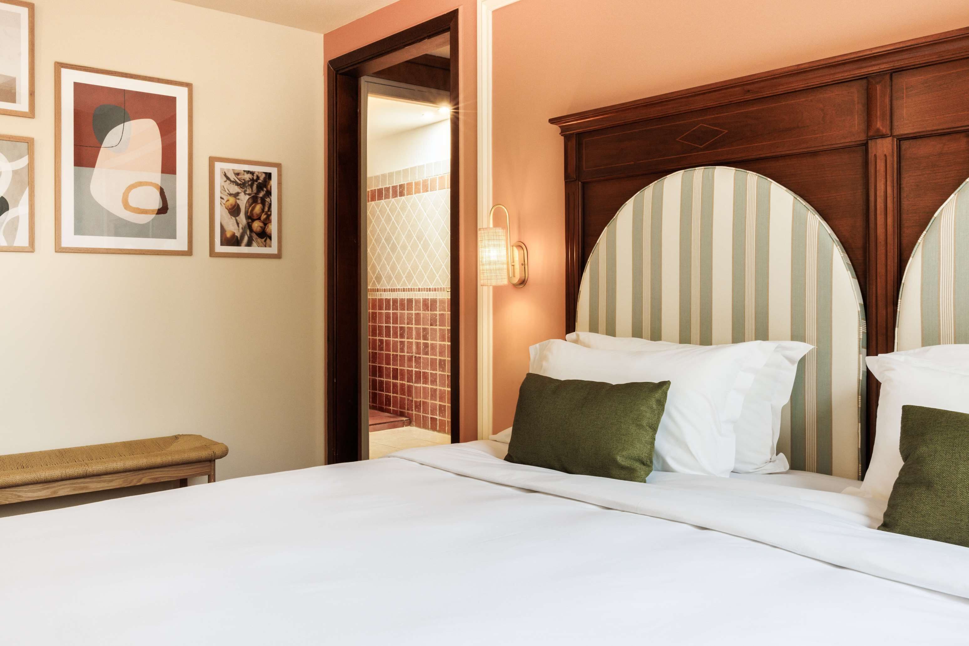 Una cama bien tendida con sábanas blancas y almohadas verdes, un banco de madera y una pared con cuadros.