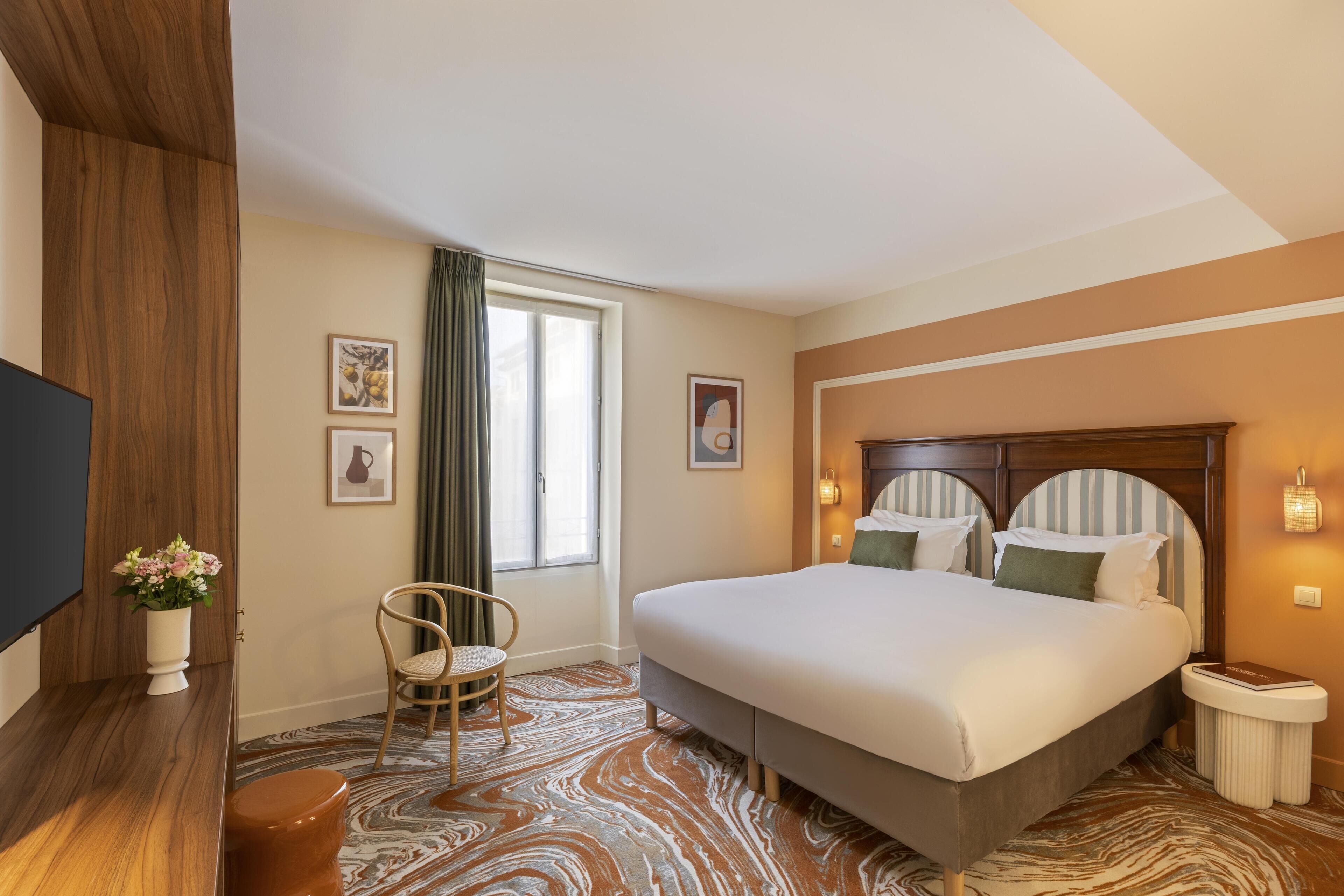 Ein Hotelzimmer mit einem großen Bett, einem Holzkopfstück, einem Nachttisch, einem Sessel, einem Fernseher und einem Fenster mit Vorhängen.