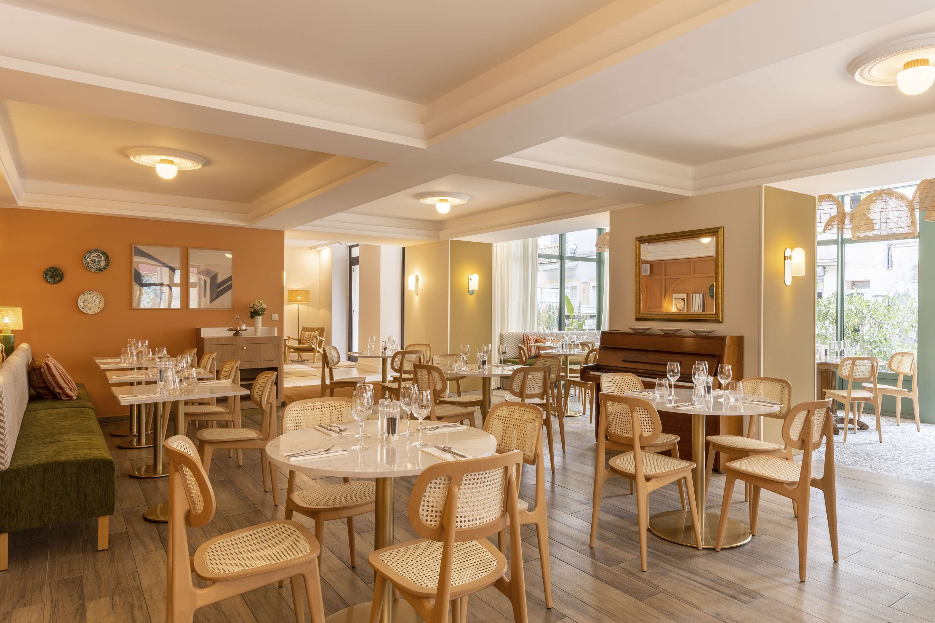 A spacious dining area with round tables and wicker chairs, large windows, and a piano in the background.