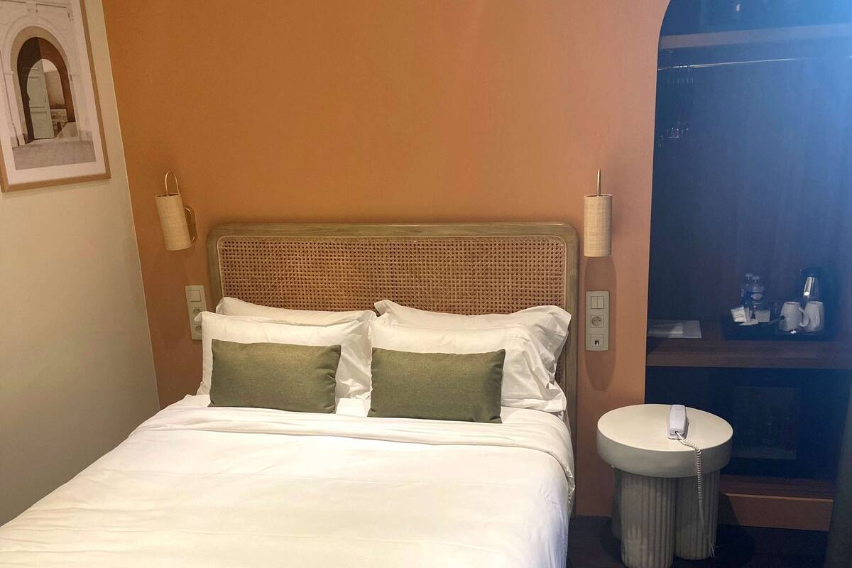 Ein ordentlich gemachtes Bett mit weißer Bettwäsche und zwei grünen Kissen, ein runder Nachttisch mit einer Lampe und ein kleiner runder Spiegel.