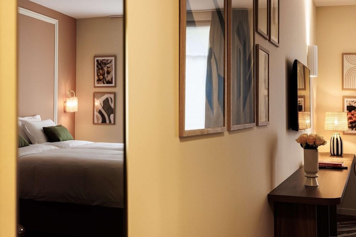 A hotel room with a bed, wall-mounted artwork, a desk with a vase, and a mirror.