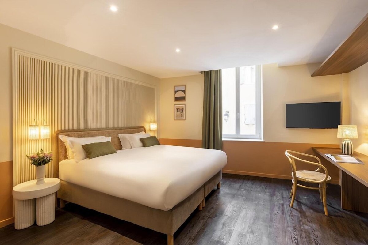 A hotel room with a bed, bedside table, lamp, chair, TV, and a window with curtains.