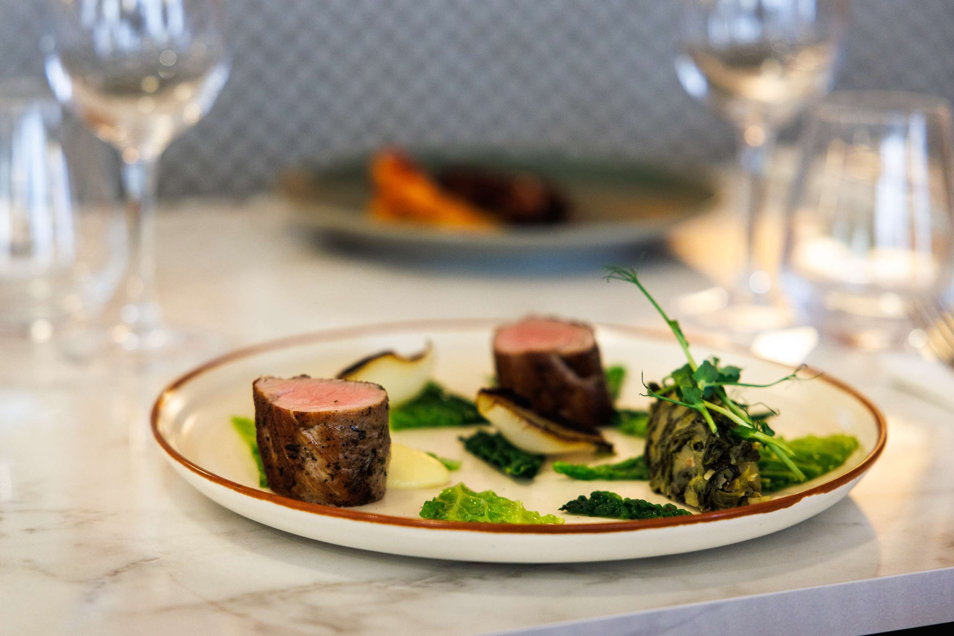 A plate of food with two pieces of meat and greens on a white plate with a brown rim.