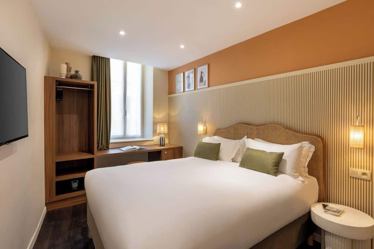 Ein Hotelzimmer mit einem großen Bett, einem Holzschrank, einem Schreibtisch mit Lampe sowie einem an der Wand befestigten Fernseher.