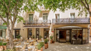 An outdoor cafe area with wicker furniture and potted plants, a covered seating area, and a building with a balcony. - Grand Hôtel d’Orange, BW Signature Collection (Orange)