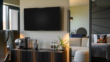 A modern hotel room with a flat-screen TV mounted on the wall, a coffee machine, a glass of water, and a potted plant on a wooden console table.