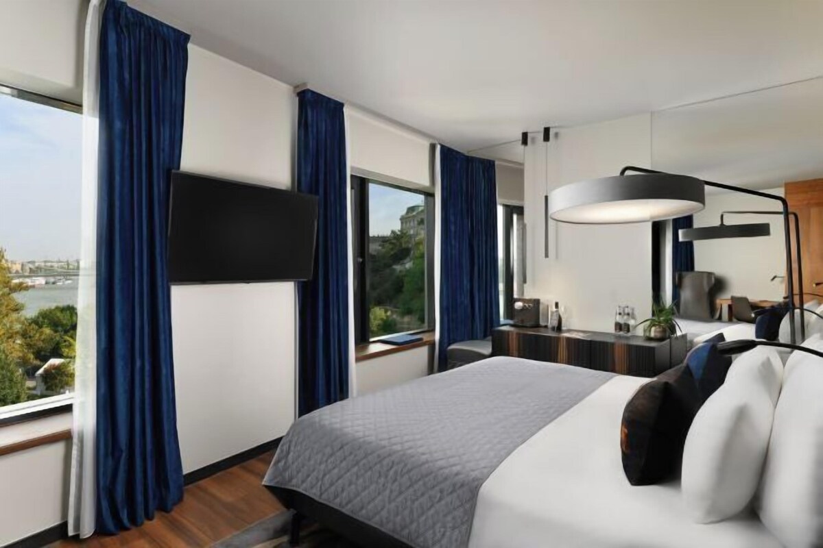 Habitación de hotel moderna con una cama grande, un escritorio, una silla, un televisor y vistas a un paisaje urbano.