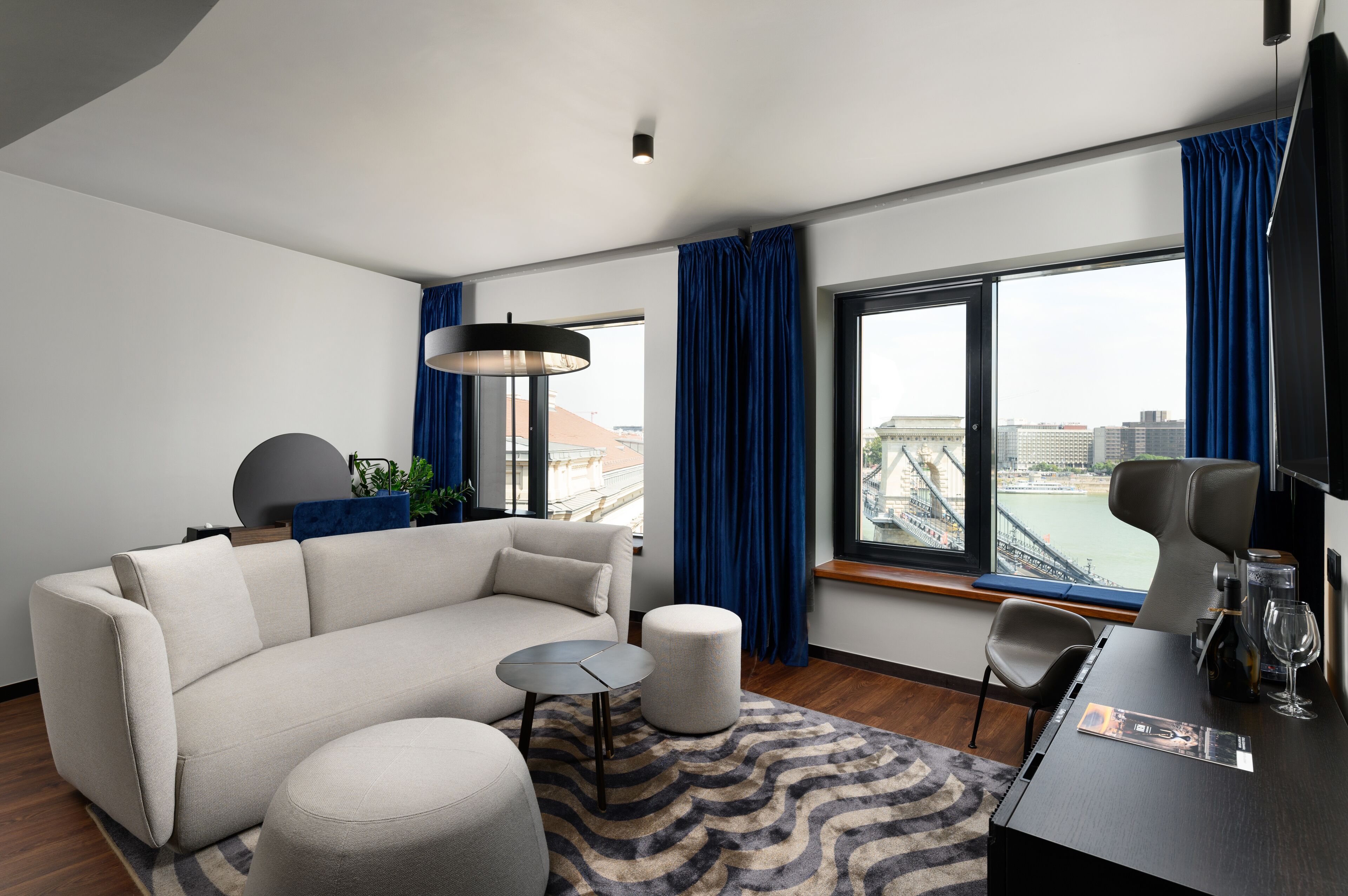 A modern hotel room with a sofa, ottoman, small table, and a view of a cityscape through large windows.