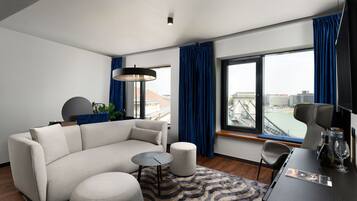 A modern hotel room with a sofa, ottoman, small table, and a view of a cityscape through large windows.