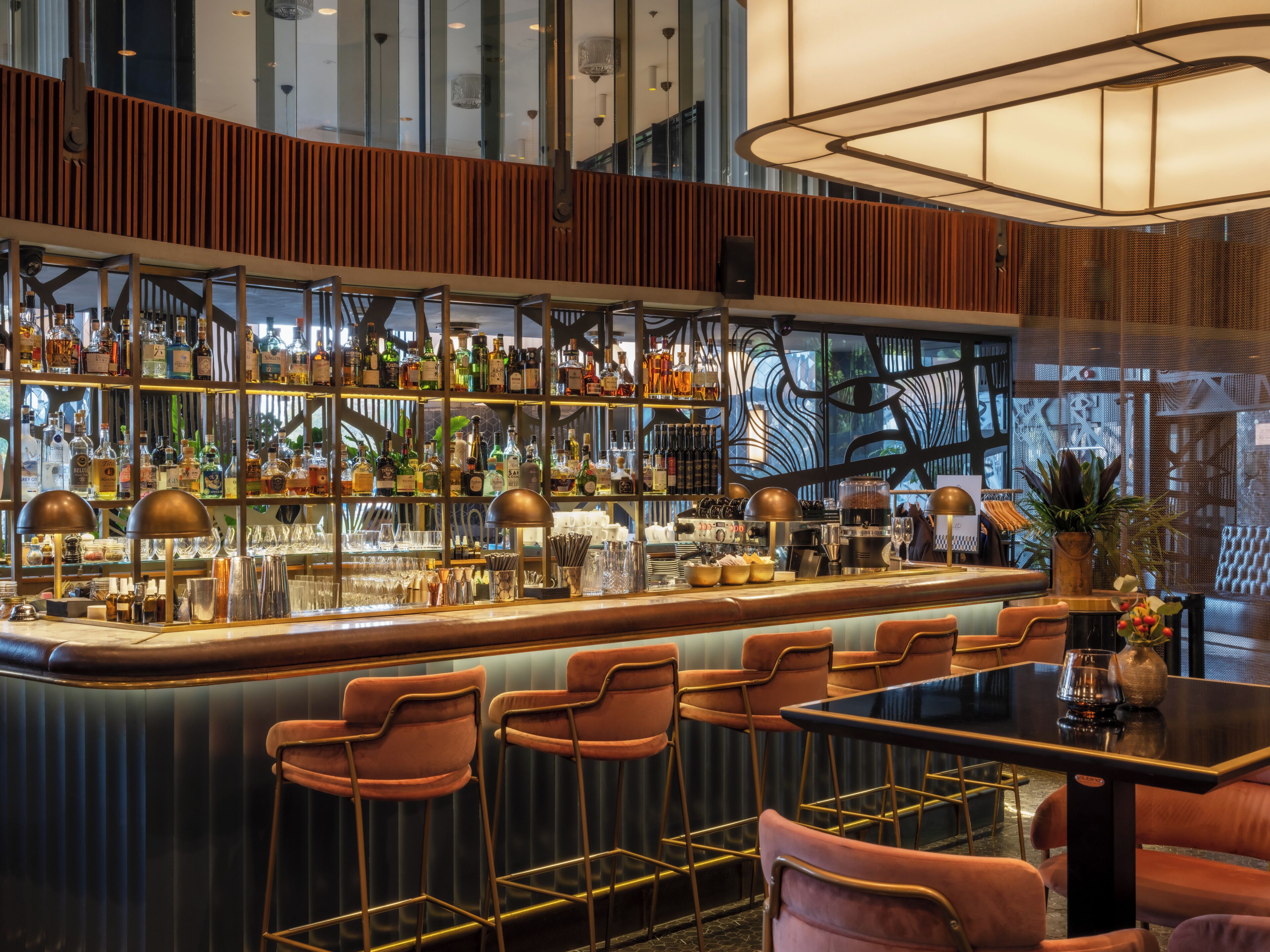 Un área de bar con una barra alta, varias estanterías llenas de botellas y una variedad de utensilios de bar. Hay taburetes de bar y una mesa con sillas.