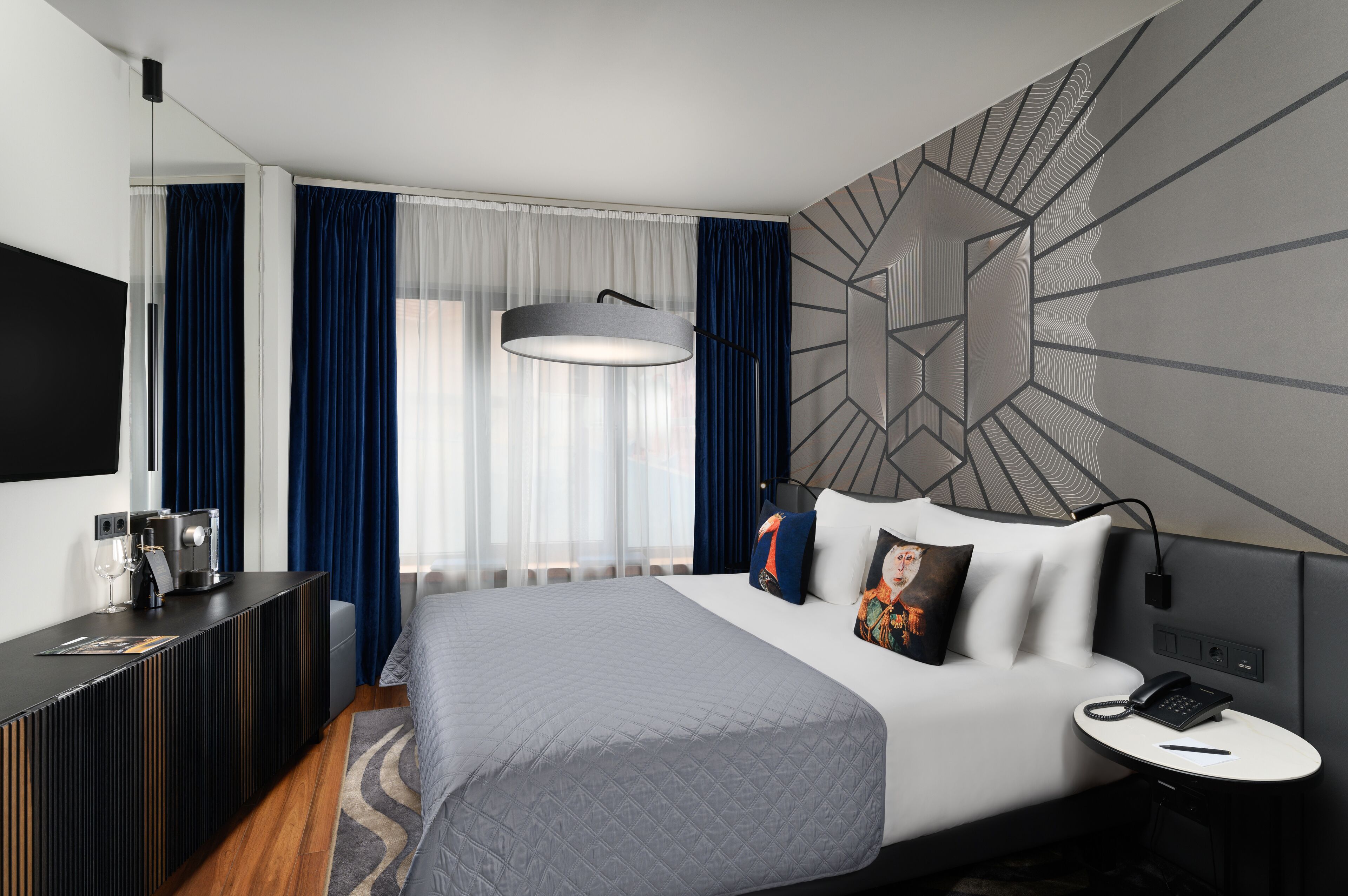 A modern hotel room with a large bed, a bedside table with a phone, a coffee machine, and a flat-screen TV.