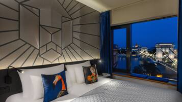 Una habitación de hotel moderna con una cama grande, diseño geométrico en la pared y vistas a un paisaje urbano al atardecer.