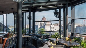 Eine moderne Rooftop-Bar mit Korbmöbeln und großen Fenstern mit Blick auf ein historisches Gebäude.