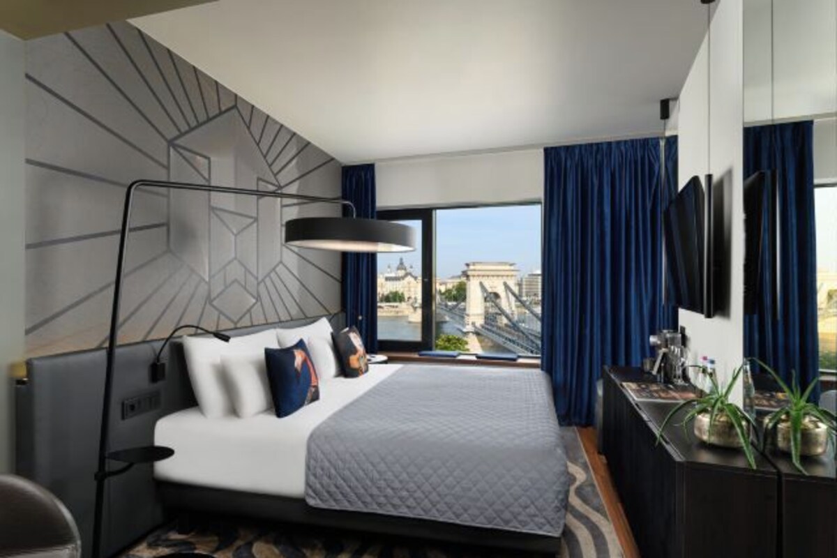 Quarto de hotel moderno com uma cama grande, uma escrivaninha com um computador, uma TV e vista para a cidade.