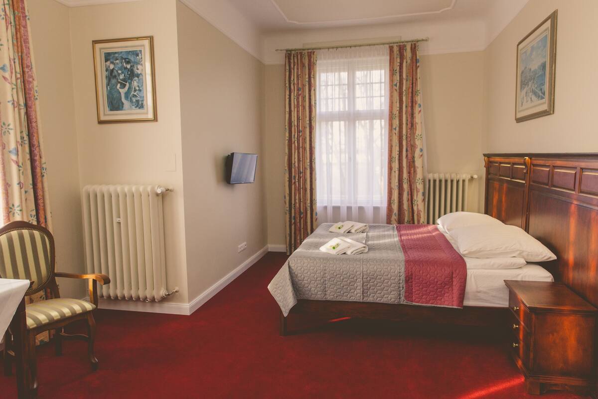 A hotel room with a bed, a chair, a table, a TV, and a radiator.