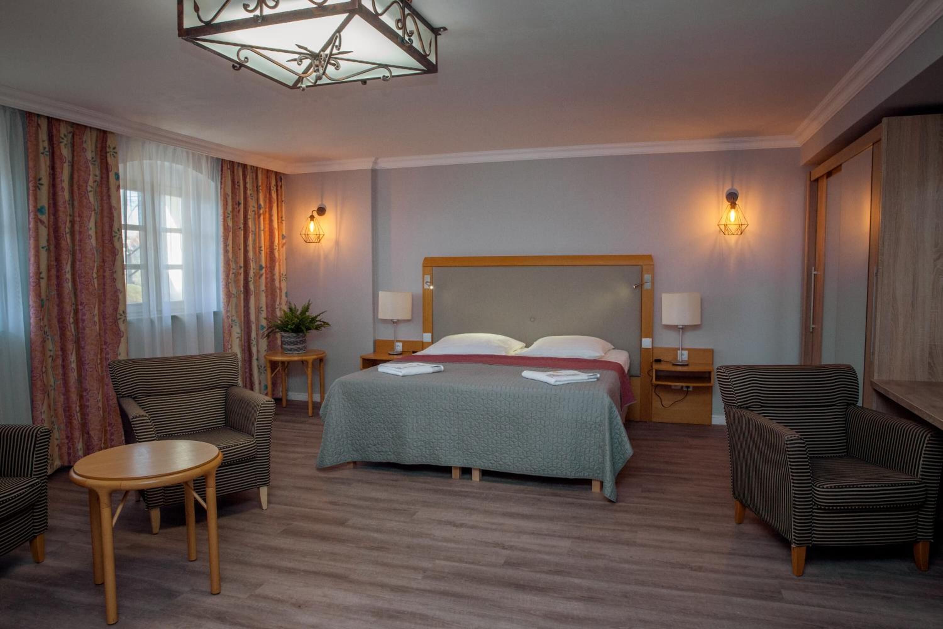 Une chambre d’hôtel avec un grand lit, deux tables de chevet, une petite table ronde, deux fauteuils et un lustre.