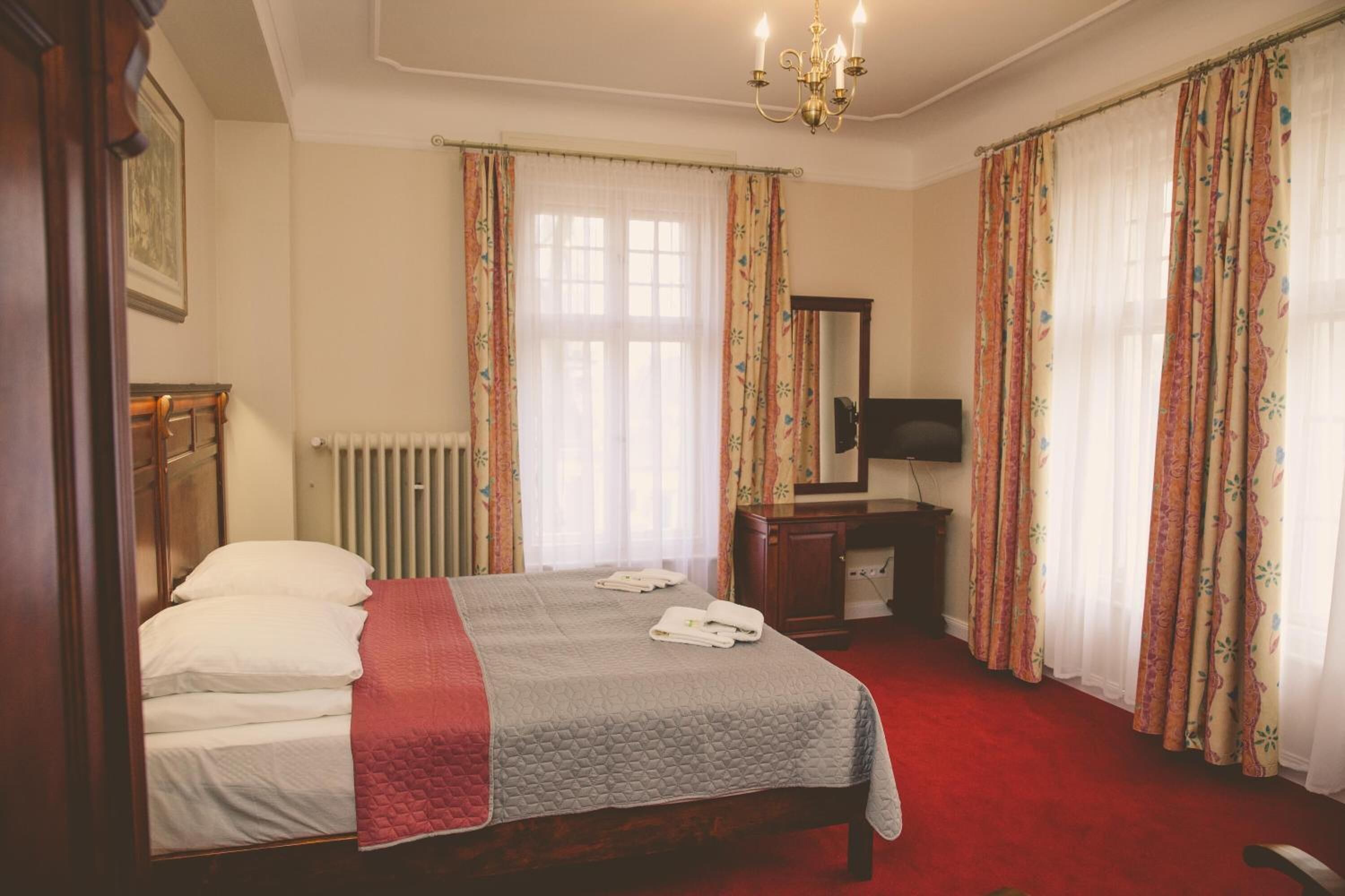 A hotel room with a bed, a desk, a chair, a radiator, and curtains.