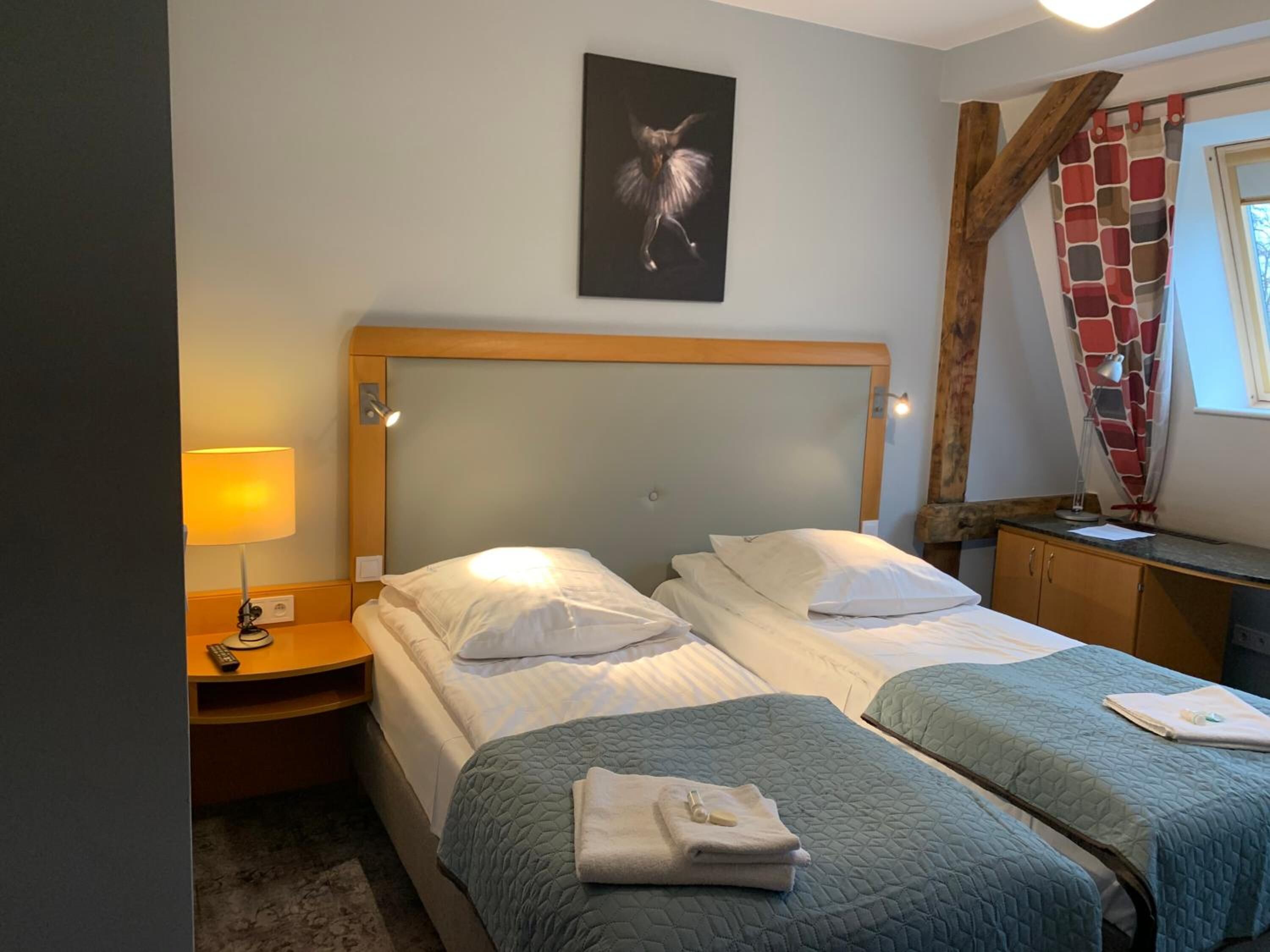 Habitación de hotel con dos camas, una mesita de noche con lámpara, cabecero de madera, un cuadro en la pared y una ventana con cortinas.