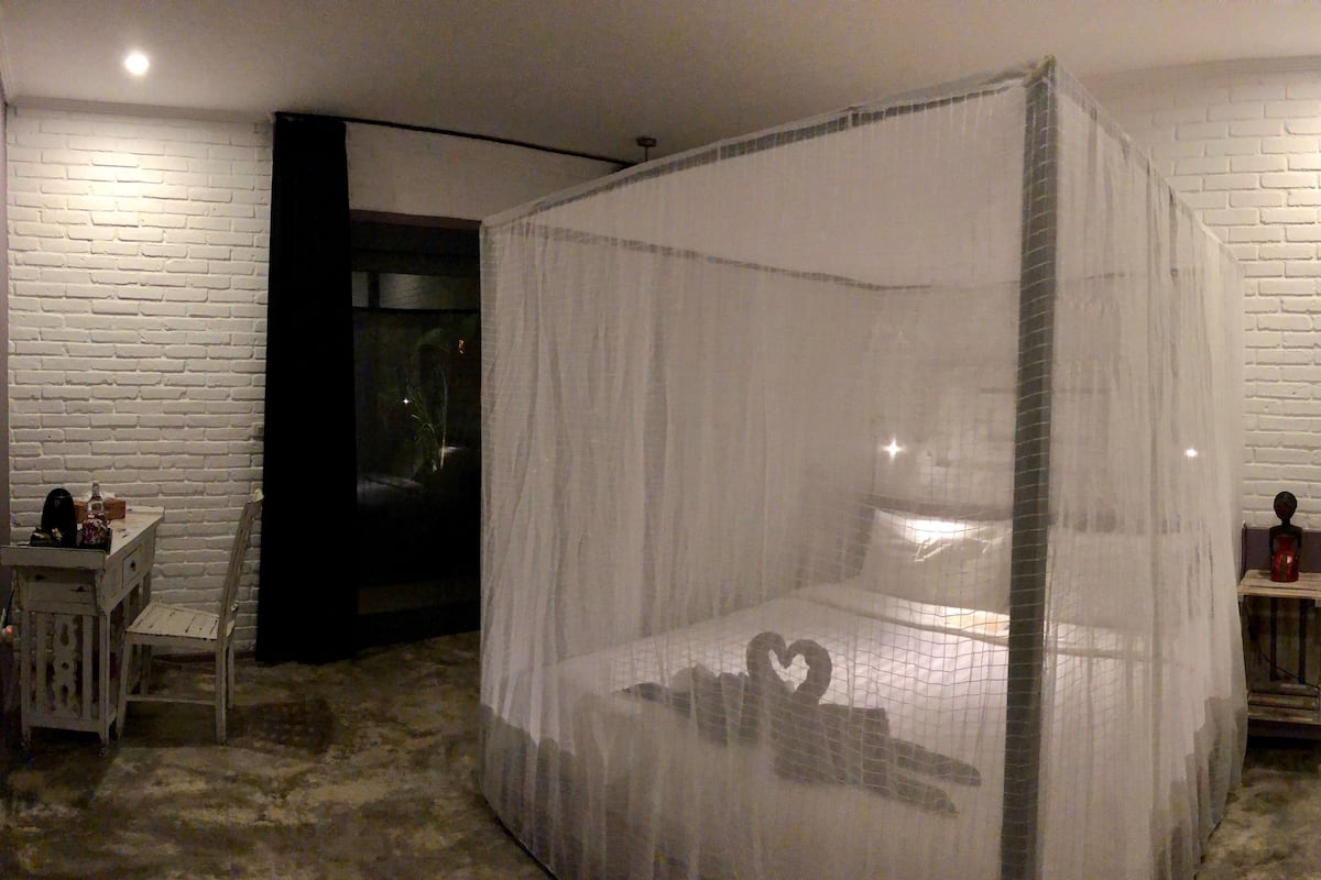 A room with a bed covered by a white canopy, a small table with a chair, and a white brick wall.