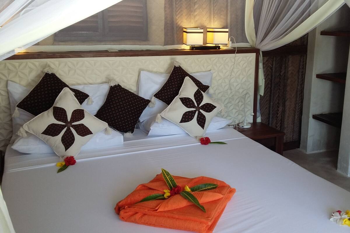 Una cama bien hecha con una toalla naranja decorativa y flores, cabecero con almohadillas estampadas, una lámpara de noche y vistas a un balcón con cortinas.