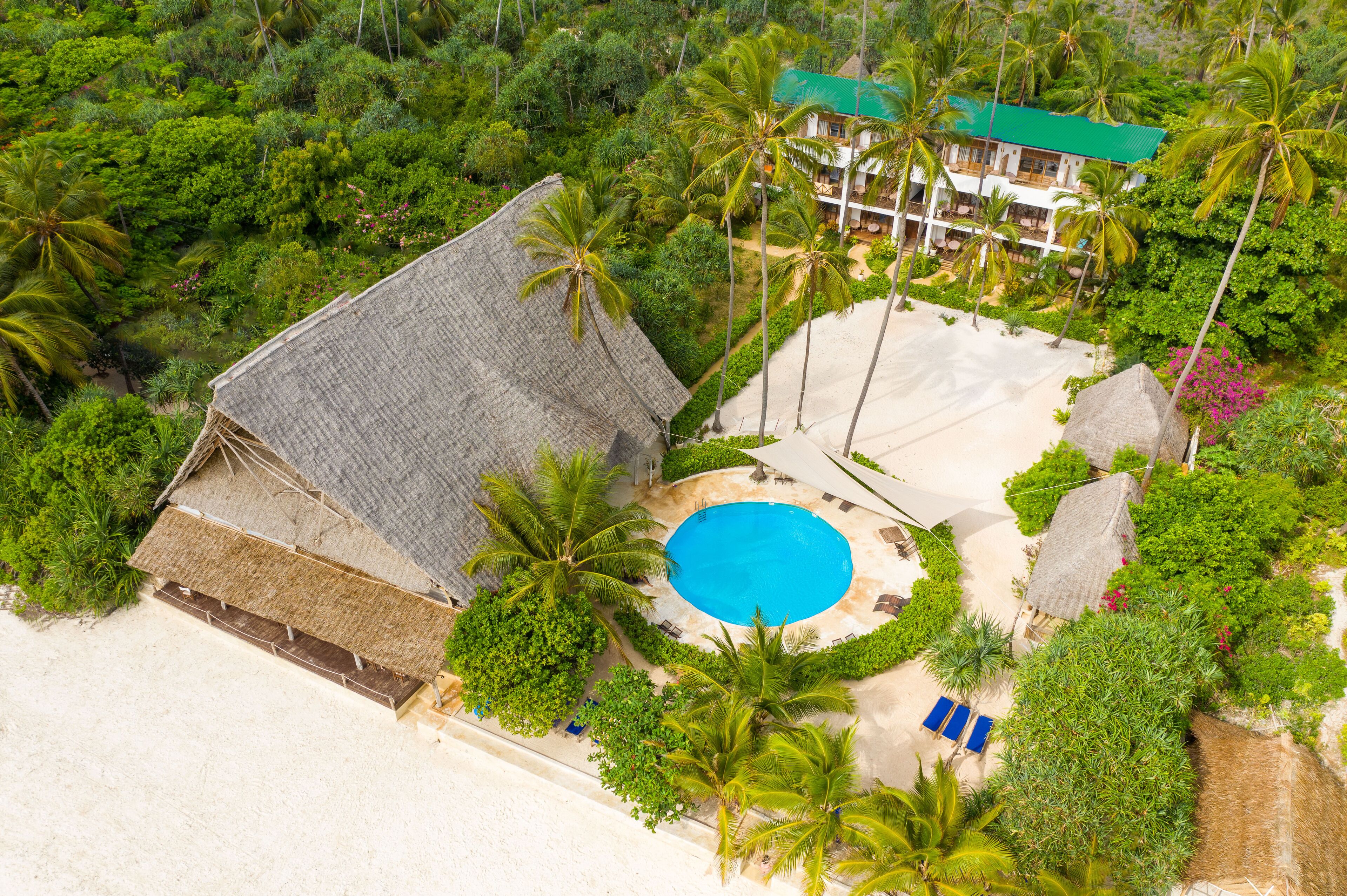 An aerial view of a resort with a swimming pool, palm trees, and a building with a green roof.