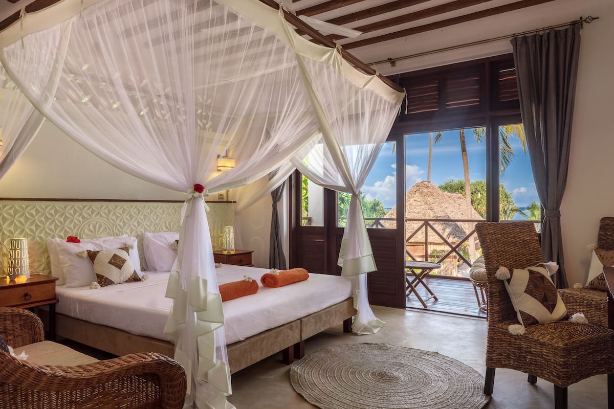 Un dormitorio con cama, dosel blanco, sillas de mimbre y vista a palmeras.