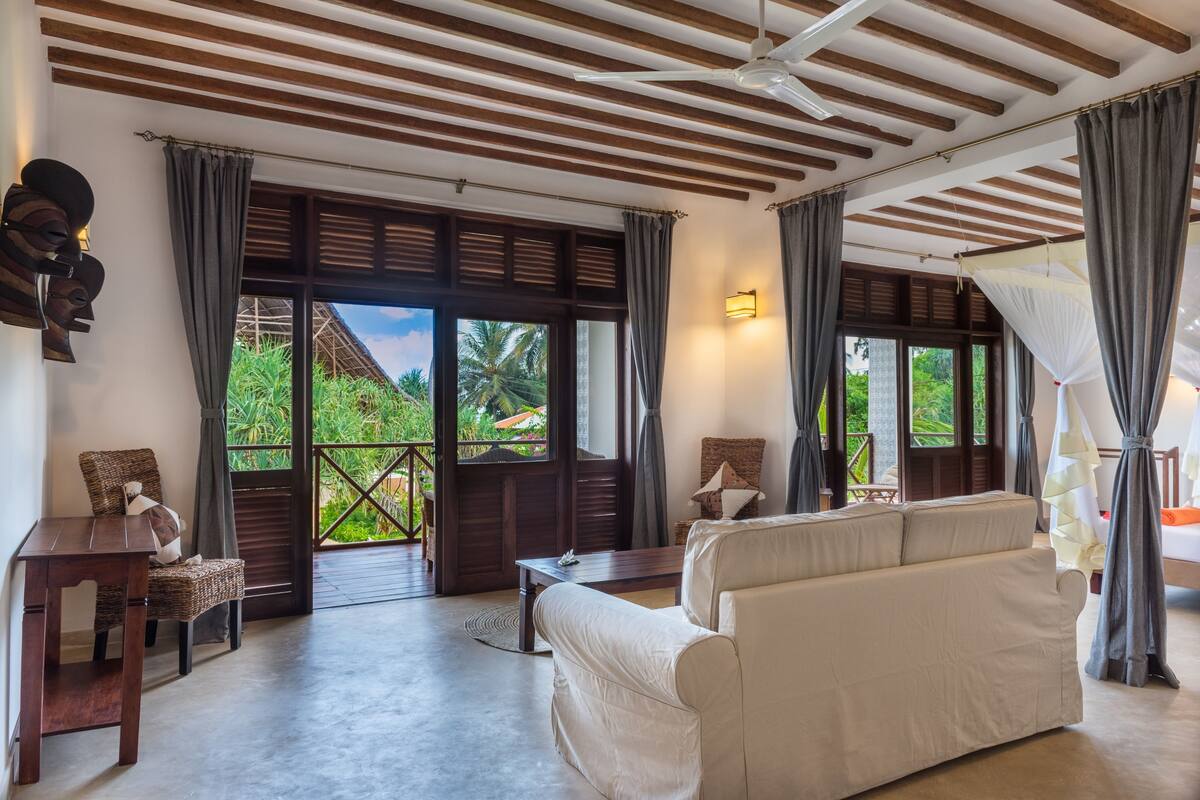 Amplia habitación con un sofá grande, una mesa de madera y una silla de mimbre. Cuenta con ventanas amplias con cortinas, un ventilador de techo y vistas a zonas verdes.