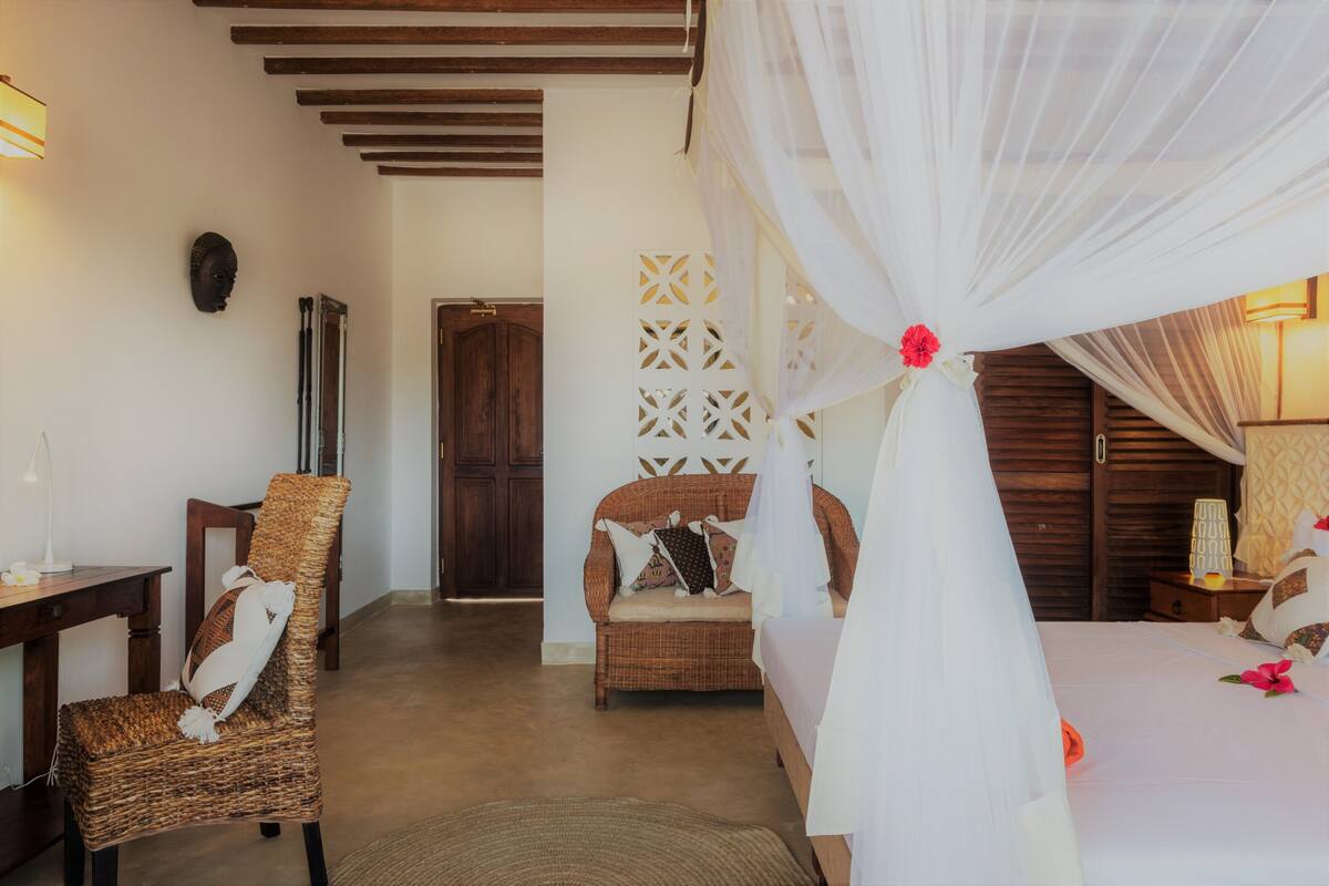 Una habitación con una cama bajo un dosel blanco, una silla de mimbre, una puerta de madera y una lámpara fijada a la pared.