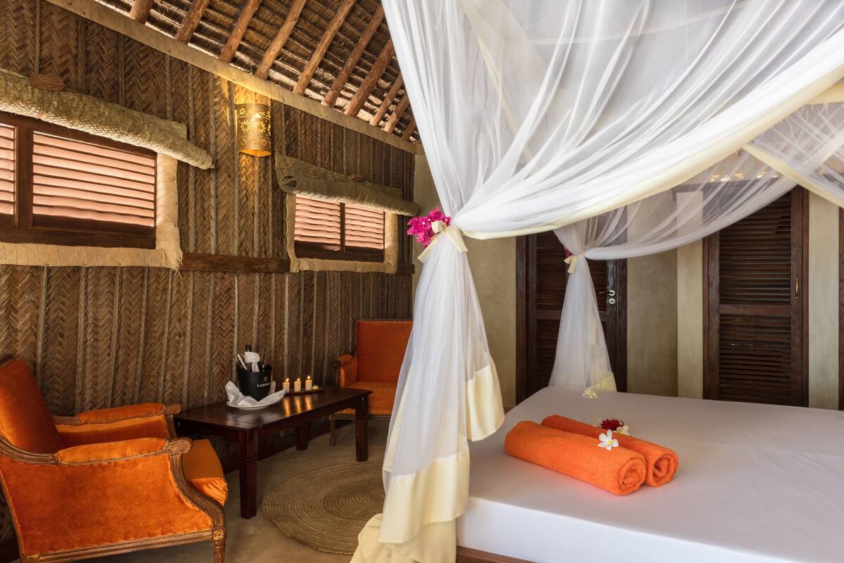 Una habitación con cama, una silla naranja, una mesa de madera y un dosel decorativo.