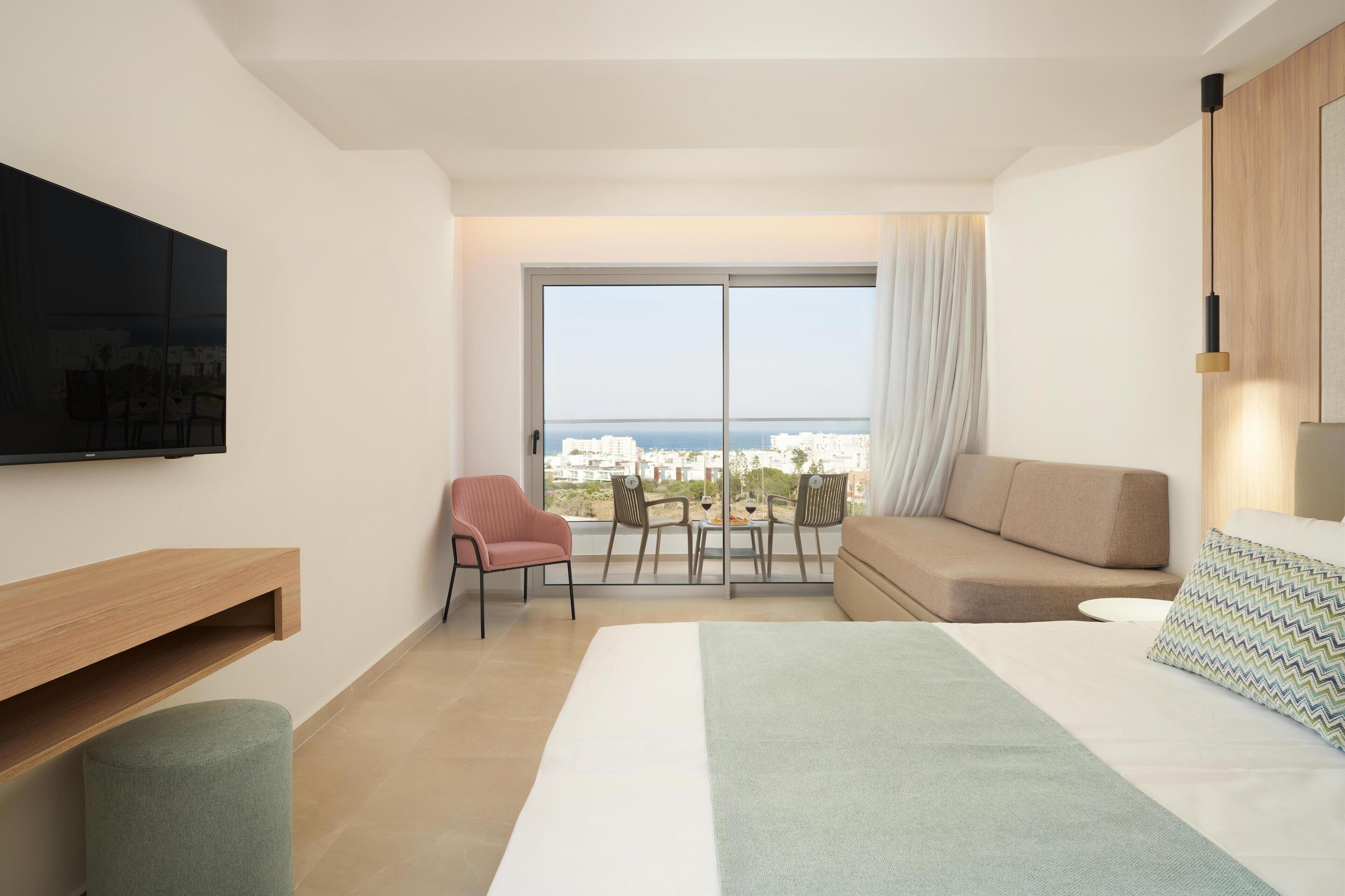Une chambre d’hôtel moderne dotée d’un grand lit, d’un téléviseur fixé au mur, d’un coin salon avec un canapé et des fauteuils, et d’une grande fenêtre offrant une vue sur la ville.