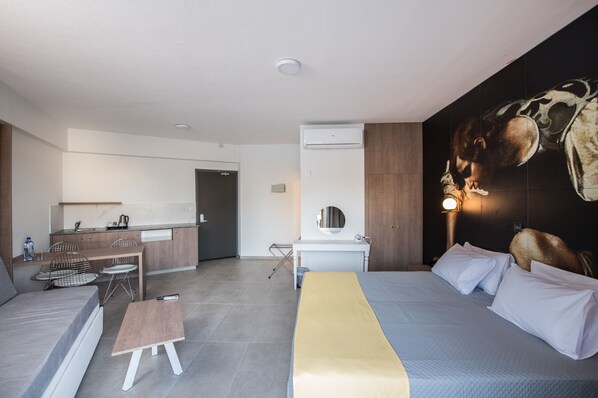 A modern hotel room with a bed, a small desk, a chair, a sofa, and a kitchen area in the background.