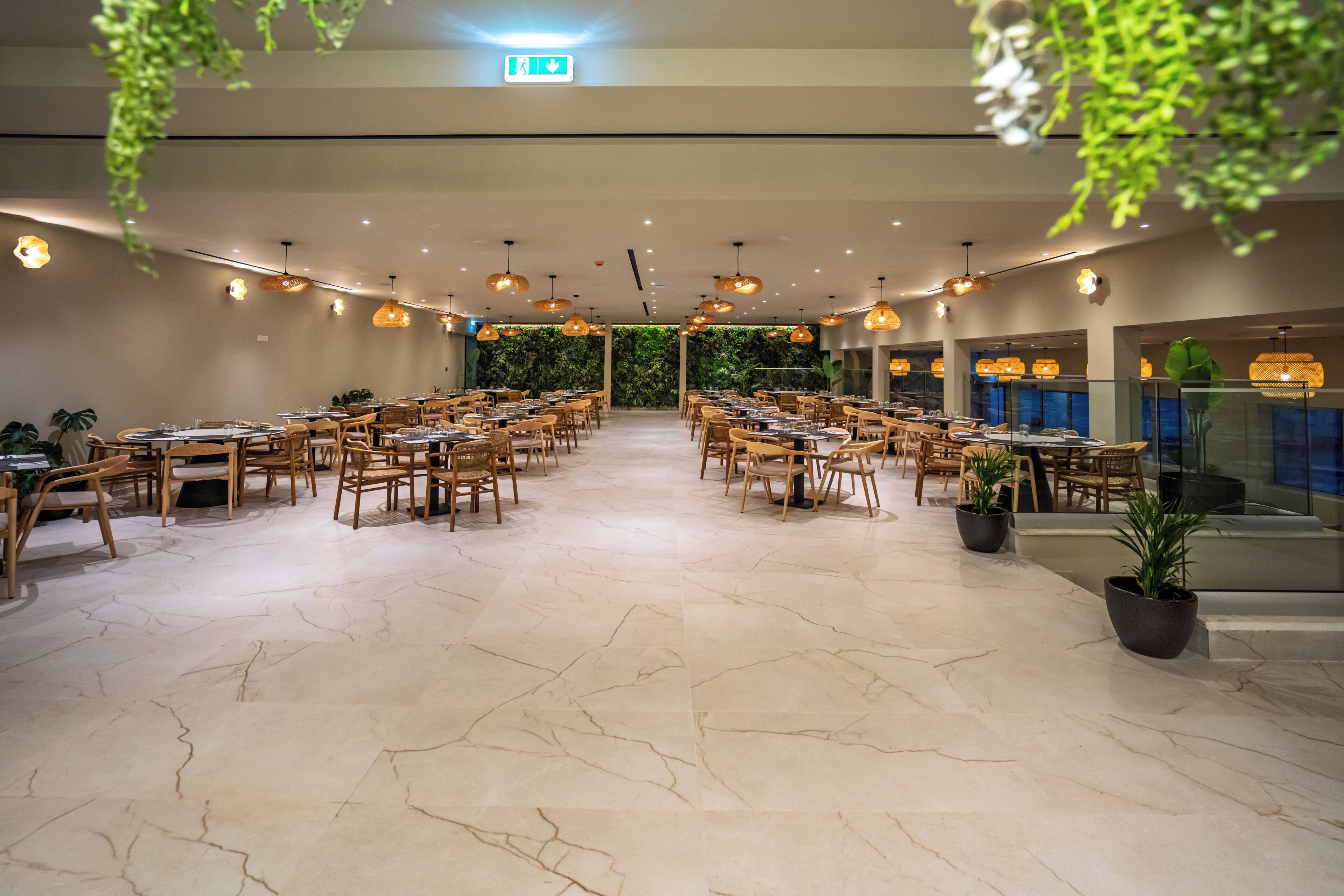 A spacious dining area with wooden tables and chairs, hanging pendant lights, and potted plants.