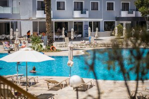 A pool area with lounge chairs and umbrellas, a palm tree, and a building with multiple windows and balconies in the background. - Narcissos Waterpark Resort (Paralimni)
