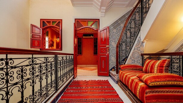 Uma escadaria com grades ornamentadas leva a um quarto com carpete vermelho e roupa de cama estampada.