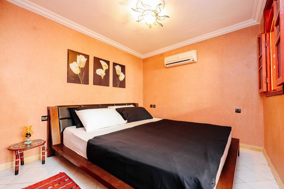 A bedroom with a bed, a small table with a lamp, and wall-mounted air conditioning.