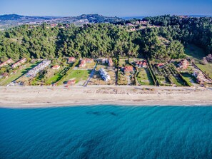 Vista aérea de un complejo turístico costero con varias villas dispuestas en filas a lo largo de la playa, rodeado de un bosque denso y colinas al fondo.