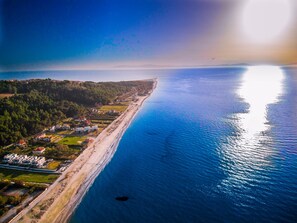 An aerial view of a coastal area with a long beach, residential buildings, and a dense forested area. - Sunset Villas (Kassandra)